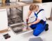 Professional technician in blue overalls repairing a built-in kitchen dishwasher using a power drill.